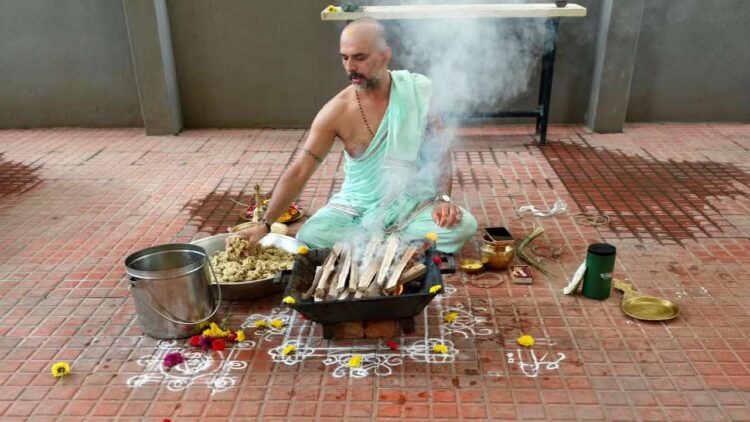 A photo showing Purohit Manjunath N Bharadwaj performing the Akhanda Maha Ganapathi Homa at the Shirdi Sai Baba Mandir premises in Bangalore. He is seated on the floor, surrounded by ceremonial items like a metal bucket, flowers, and a brass pot, while placing offerings into a square fire pit from which smoke is rising. He is wearing a light mint-green traditional attire and has a watch on his left wrist.