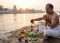 A man performing Pinda Pradana and Tarpanam rituals by a riverbank during sunrise, with a temple visible in the background.
